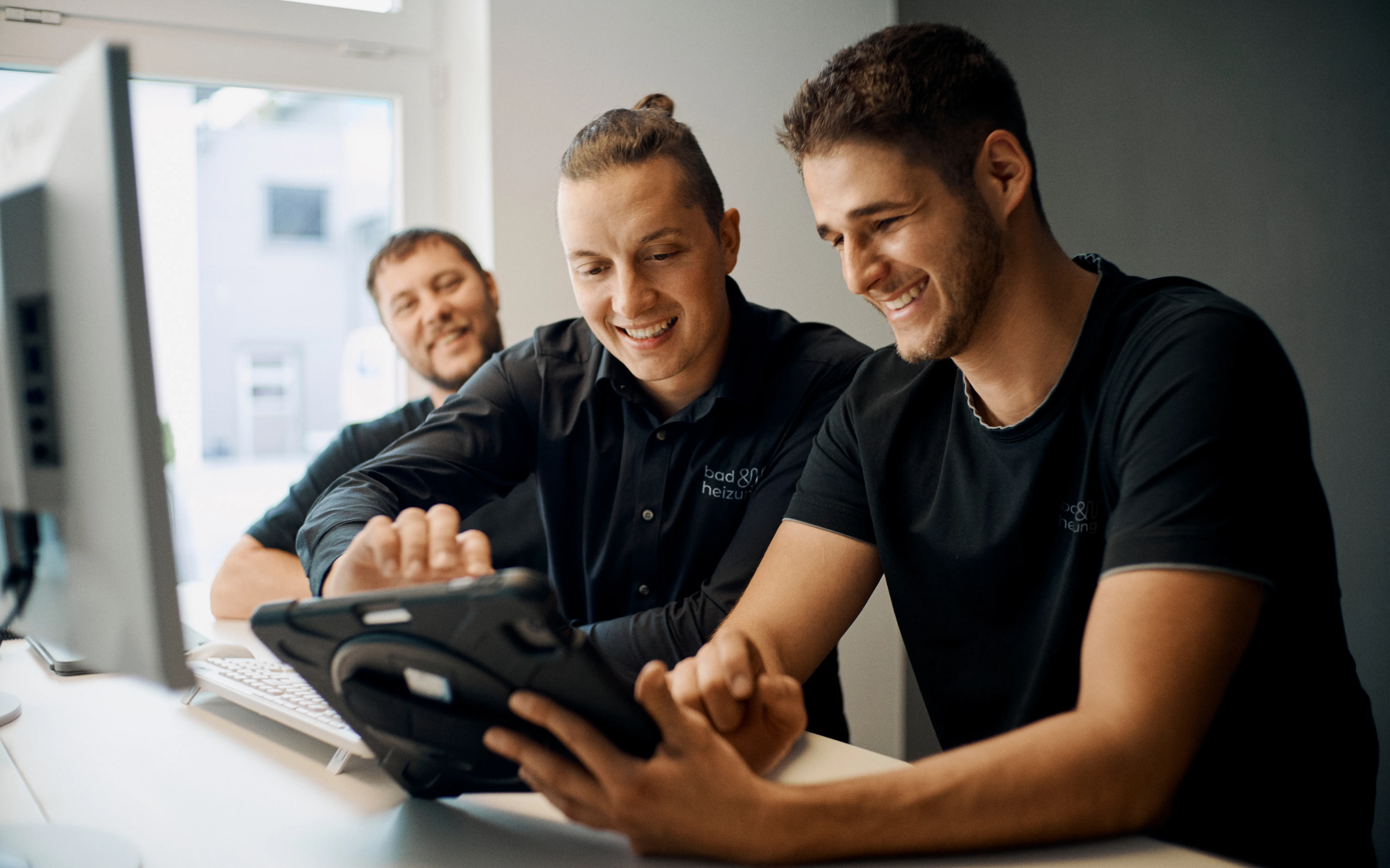 Drei Kollegen stehen an einem Schreibtisch und arbeiten gemeinsam an einem Tablet, das von einem der Männer gehalten wird. 