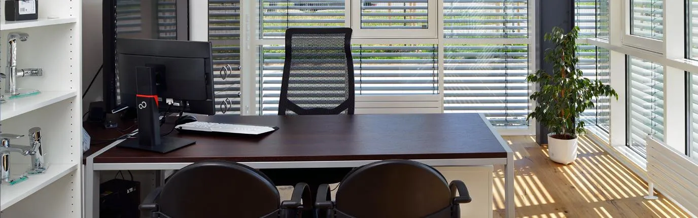 Ein modernes Büro mit einem großen Schreibtisch aus dunklem Holz, einem schwarzen Bürostuhl und einem Computerbildschirm, der zentral auf dem Tisch steht.