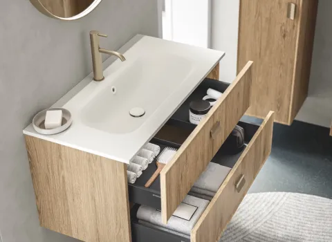 Wandhängender Waschtisch in Holzoptik mit geöffneten Schubladen und goldfarbener Armatur unter rundem Spiegel.