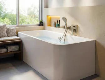 Wandstehende Badewanne mit glänzender Oberfläche und Wandarmatur. Links steht eine Bank mit Kissen und Handtüchern, dahinter ein großes Fenster mit Blick auf die gruene Natur.